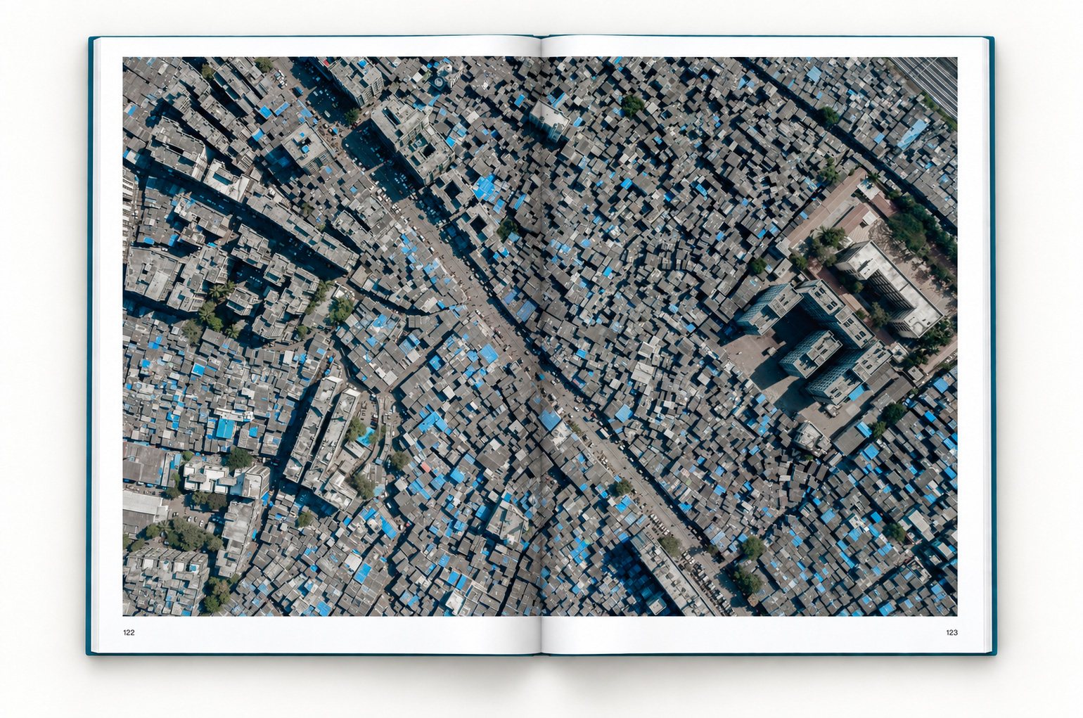 The book open at pages 122 and 123. One photograph runs across both pages: the gray concrete roofs of Dharavi, many covered in blue tarpaulins, packed along narrow lanes, with a few apartment blocks at the edge.