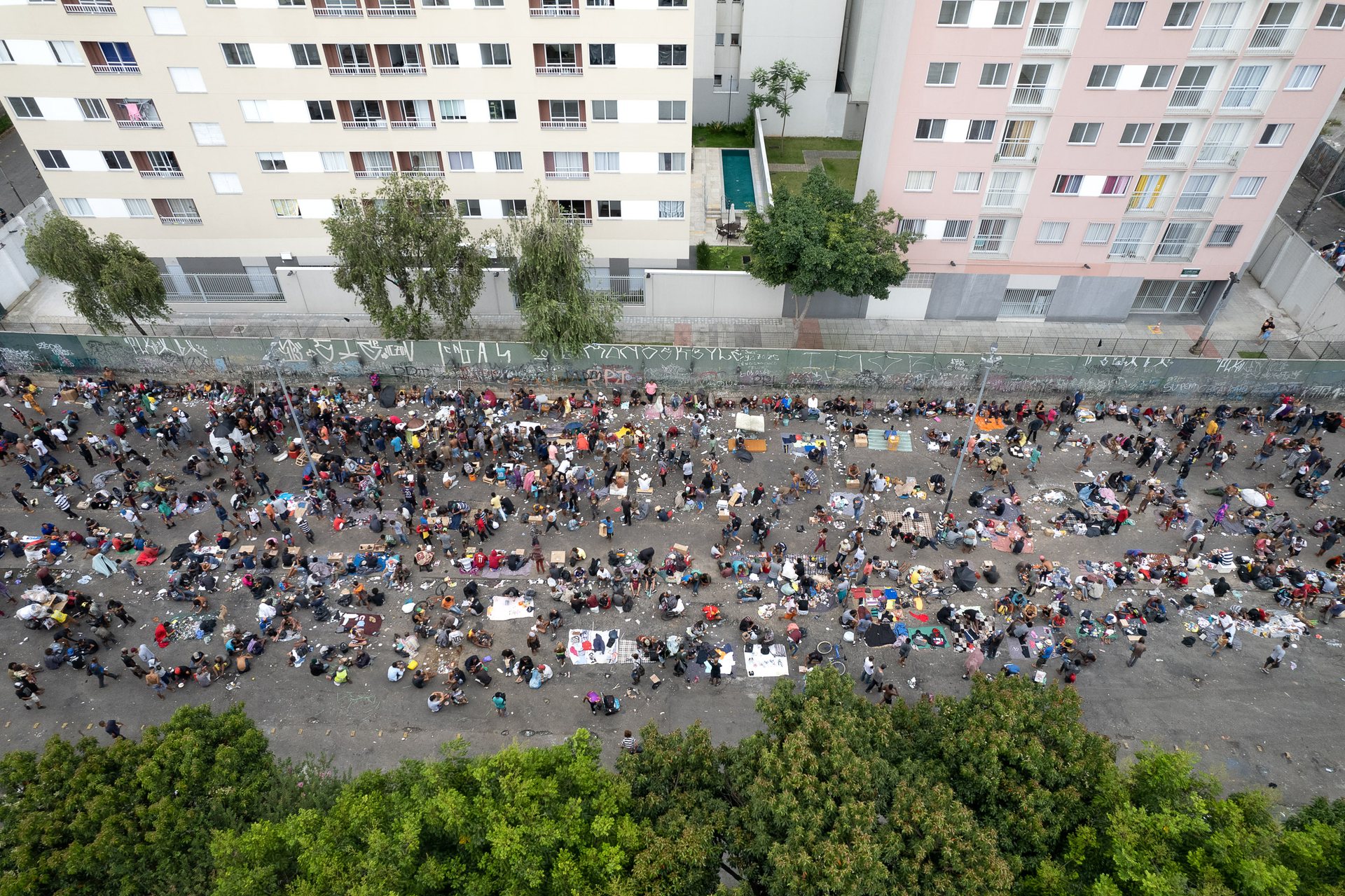 Overhead view of a large crowd gathered in São Paulo’s Cracolândia, an open-air drug zone surrounded by apartment buildings and trees.