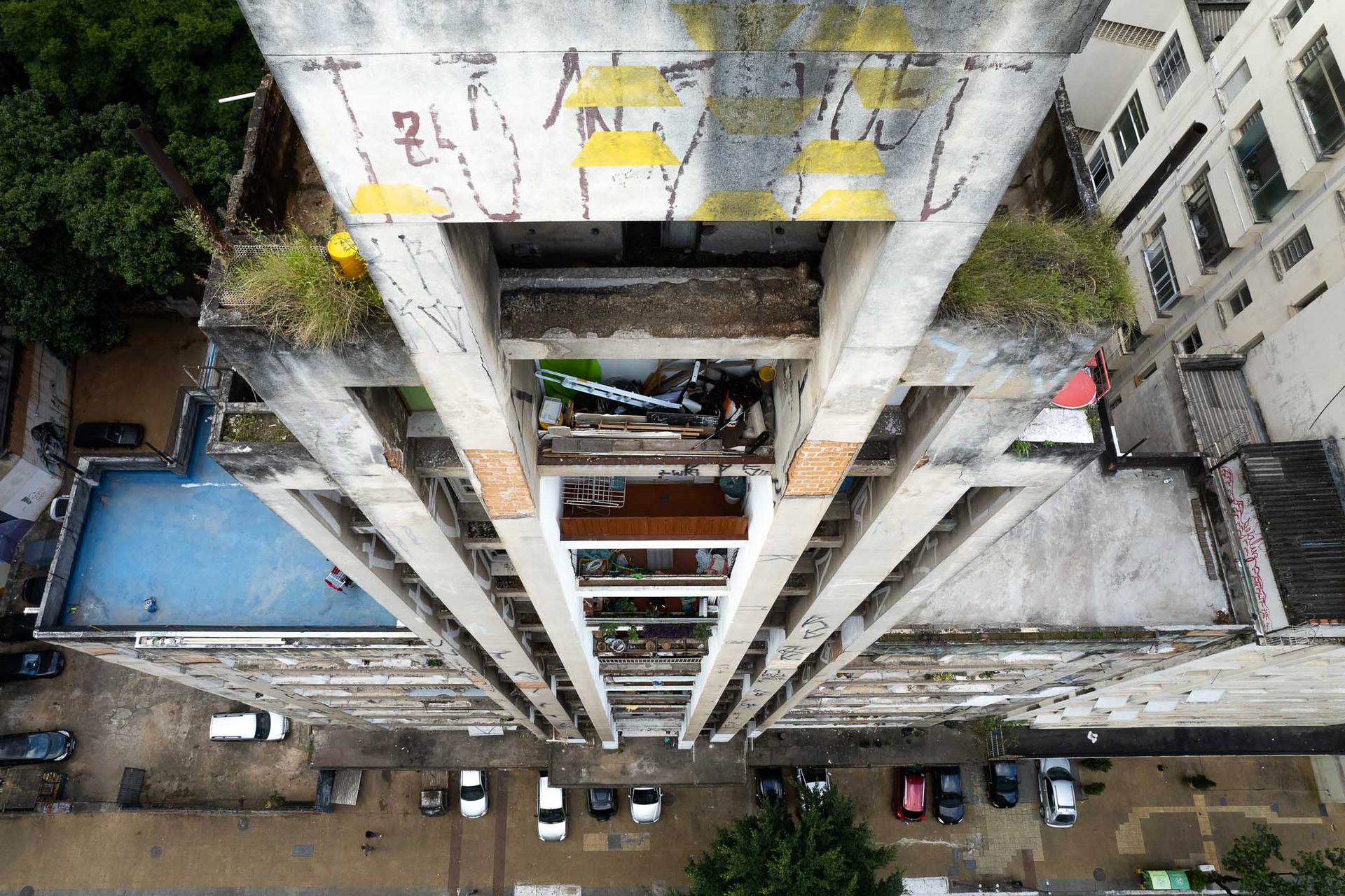 Vertigo-inducing downward aerial of Occupy 9 de Julho housing movement in central São Paulo, showing a concrete tower, rooftop vegetation, a basketball court, and the dense city below.