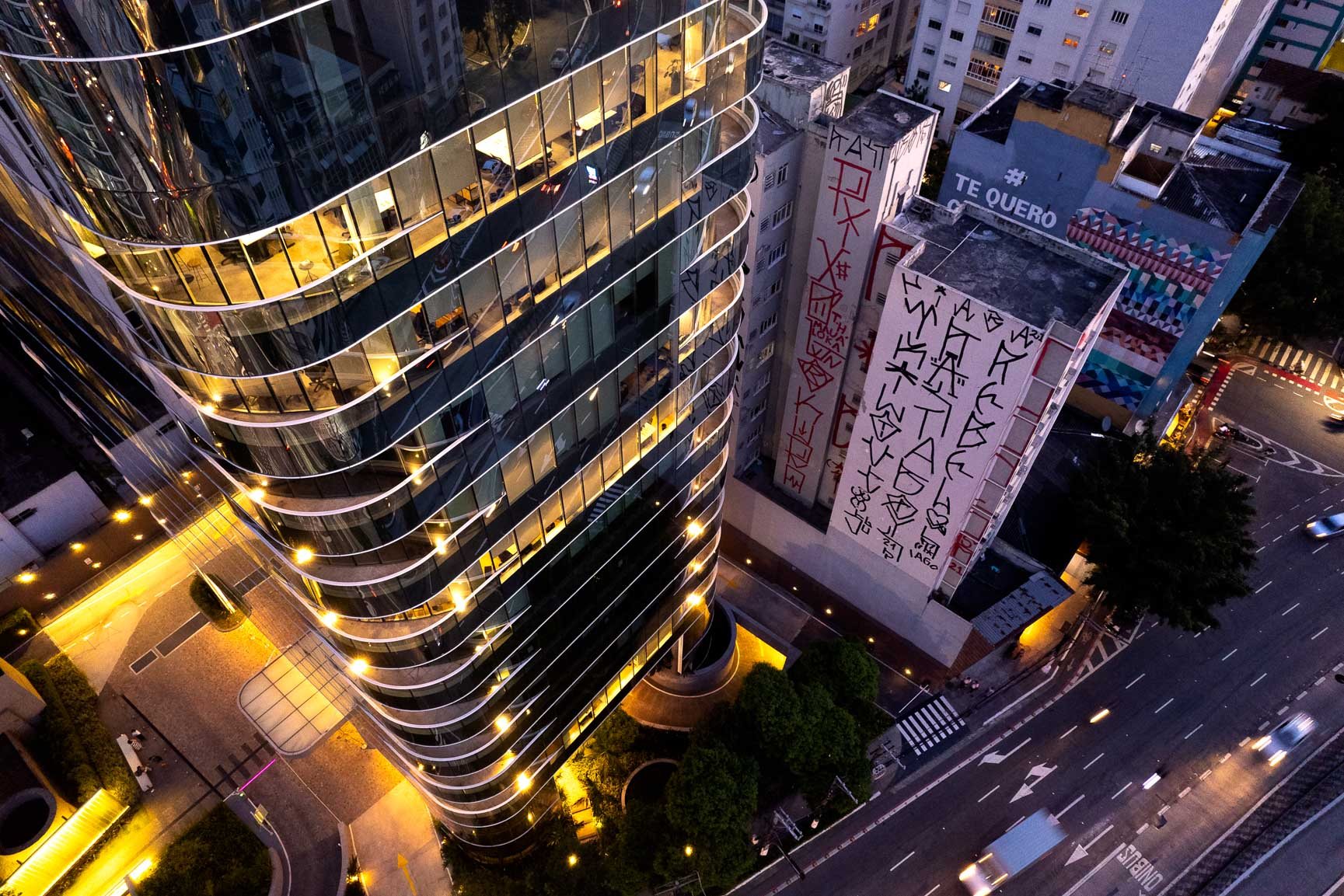 Aerial view of a modern glass building in São Paulo at night, juxtaposed with a neighboring structure covered in vertical 'pichação' graffiti, symbolizing protest culture.