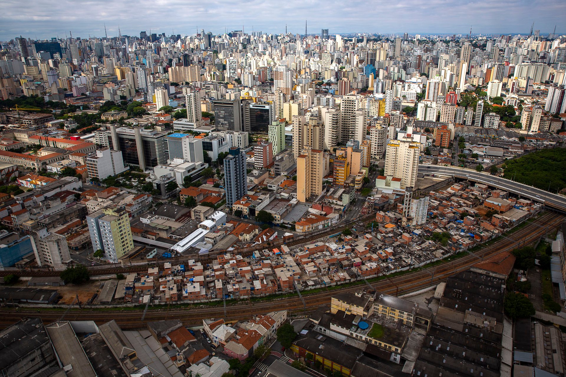 Bird’s-eye view of Moinho favela, São Paulo’s last central favela, compressed between train tracks and high-rise commercial buildings, showing extreme urban density.