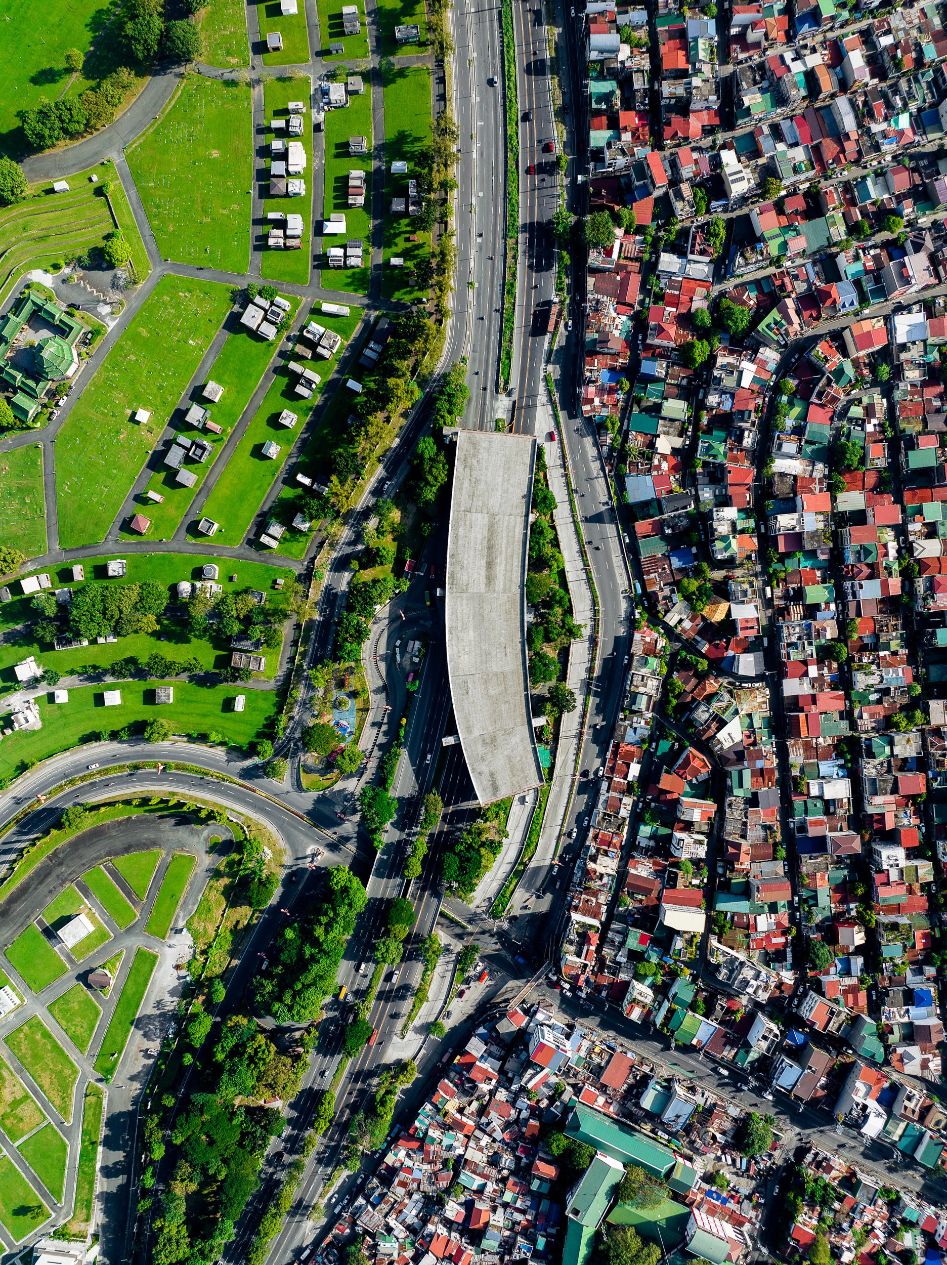 Aerial photograph of Manila showing a highway dividing a cemetery with green lawns and orderly plots from a high-density urban residential neighborhood with narrow winding streets.