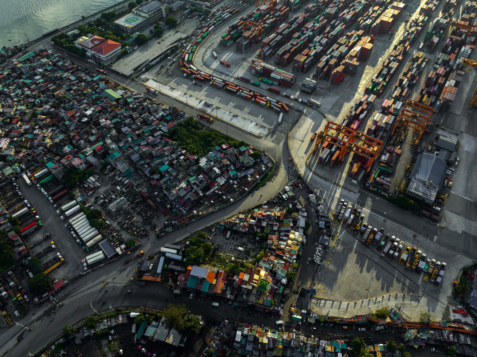 Aerial view of Manila’s port district showing stark contrast between dense informal settlements and an industrial shipping yard filled with shipping containers and cranes, highlighting spatial inequality in the Philippines.