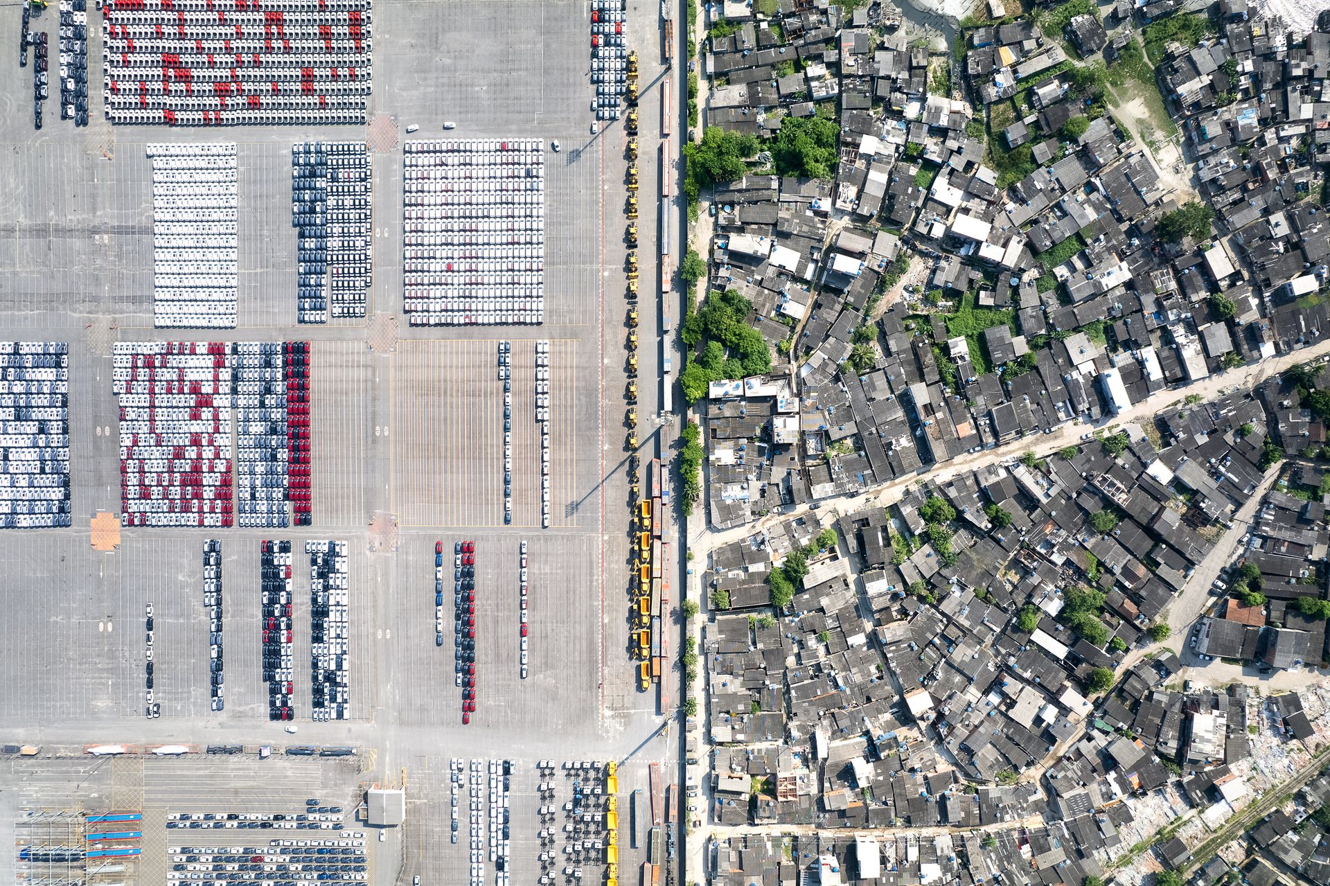 Drone photo showing a stark divide between the orderly rows of cars in the Santos port storage yard and the irregular, densely packed rooftops of a nearby favela, separated by a narrow road.