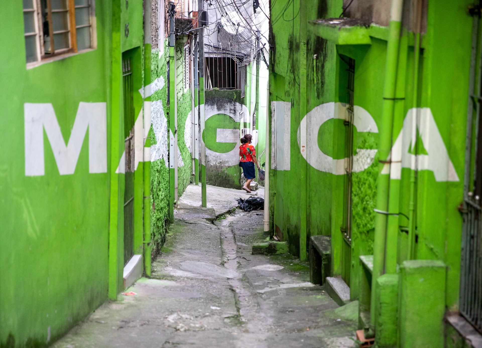 Green alleyway in the Vila Brasilândia favela, São Paulo, with the word "MÁGICA" painted across walls in a perspective-based mural by Boa Mistura.